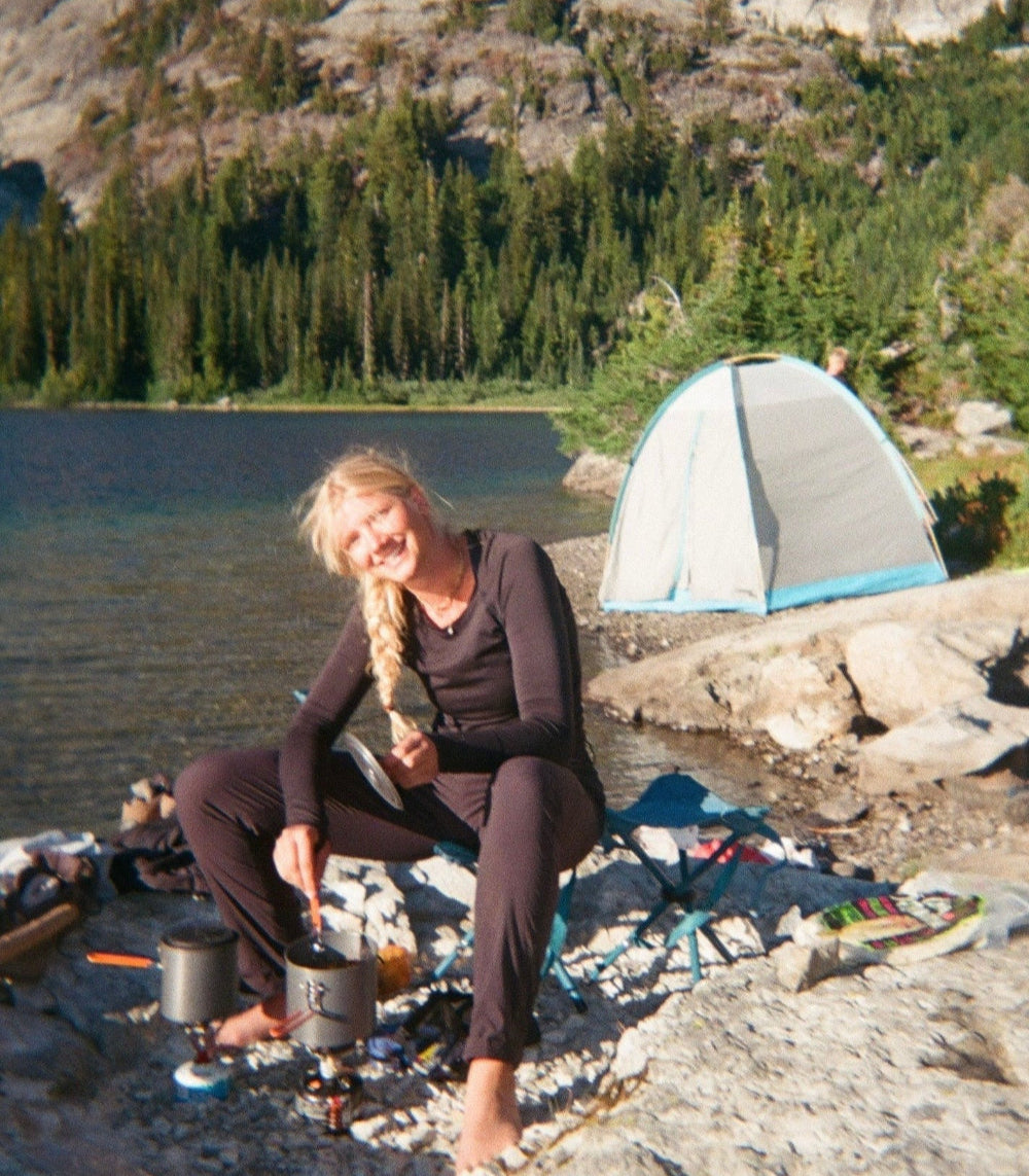 a blonde girl sitting in a chair cooking a meal on a portable stove. background is of a lake in the mountains and a tent on the bank.
