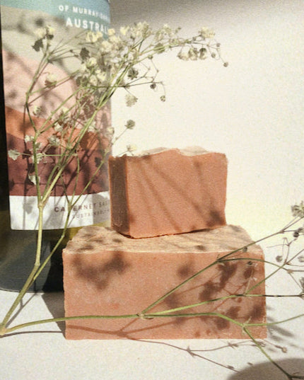 light pink soap with a small soap stacked on top. there are little white flowers surrounding it with a wine bottle in the background