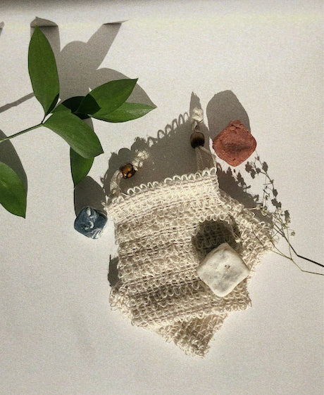 soap saver sisal bags laying down and stacked on one another with 3 little squares of soap scattered around it. One on the top right corner, one on the top of the left side and one on top of the sisal bag at the bottom right. There is a stem of leaves right above the bags. 